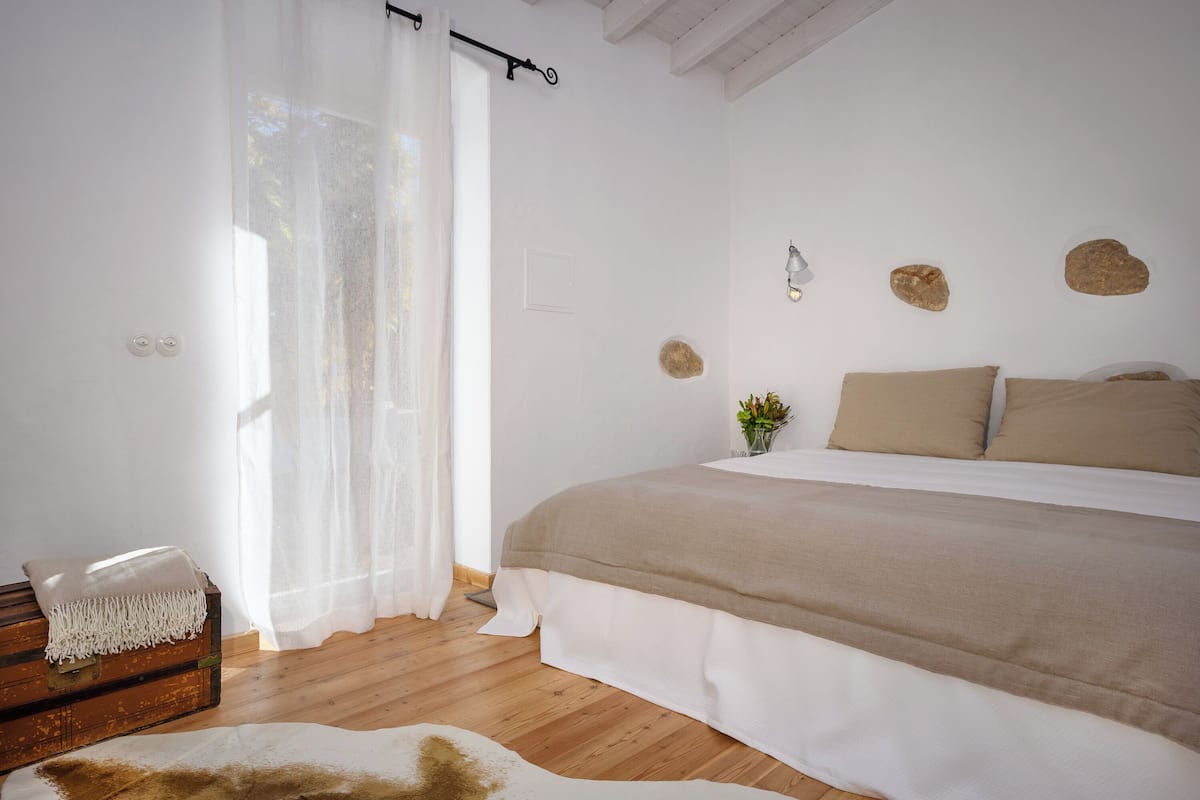 A bedroom with a wooden floor, a bed with beige bedding, a white rug, a wooden chest, and a sheer curtain.