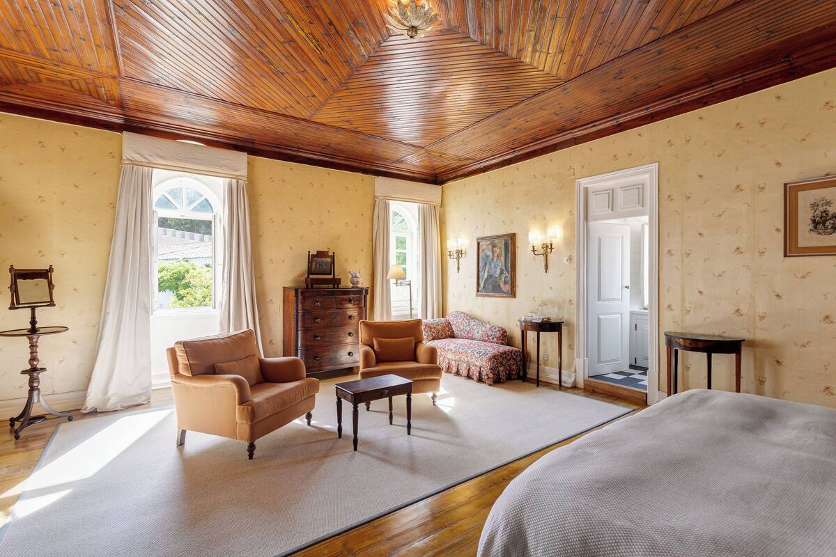A spacious living room with a wooden ceiling, large windows, a sofa, a coffee table, and a bed.