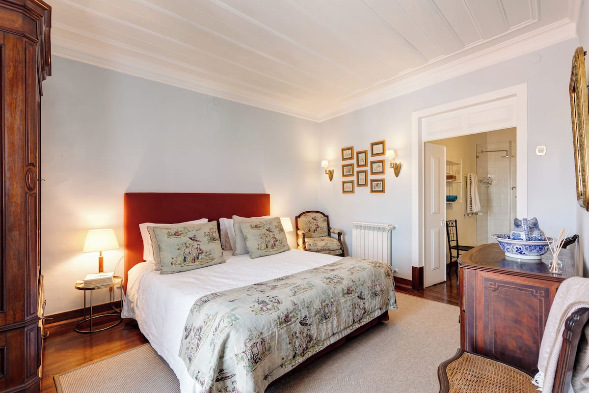 A bedroom with a large bed, a wooden wardrobe, a small table with a lamp, and a chair.