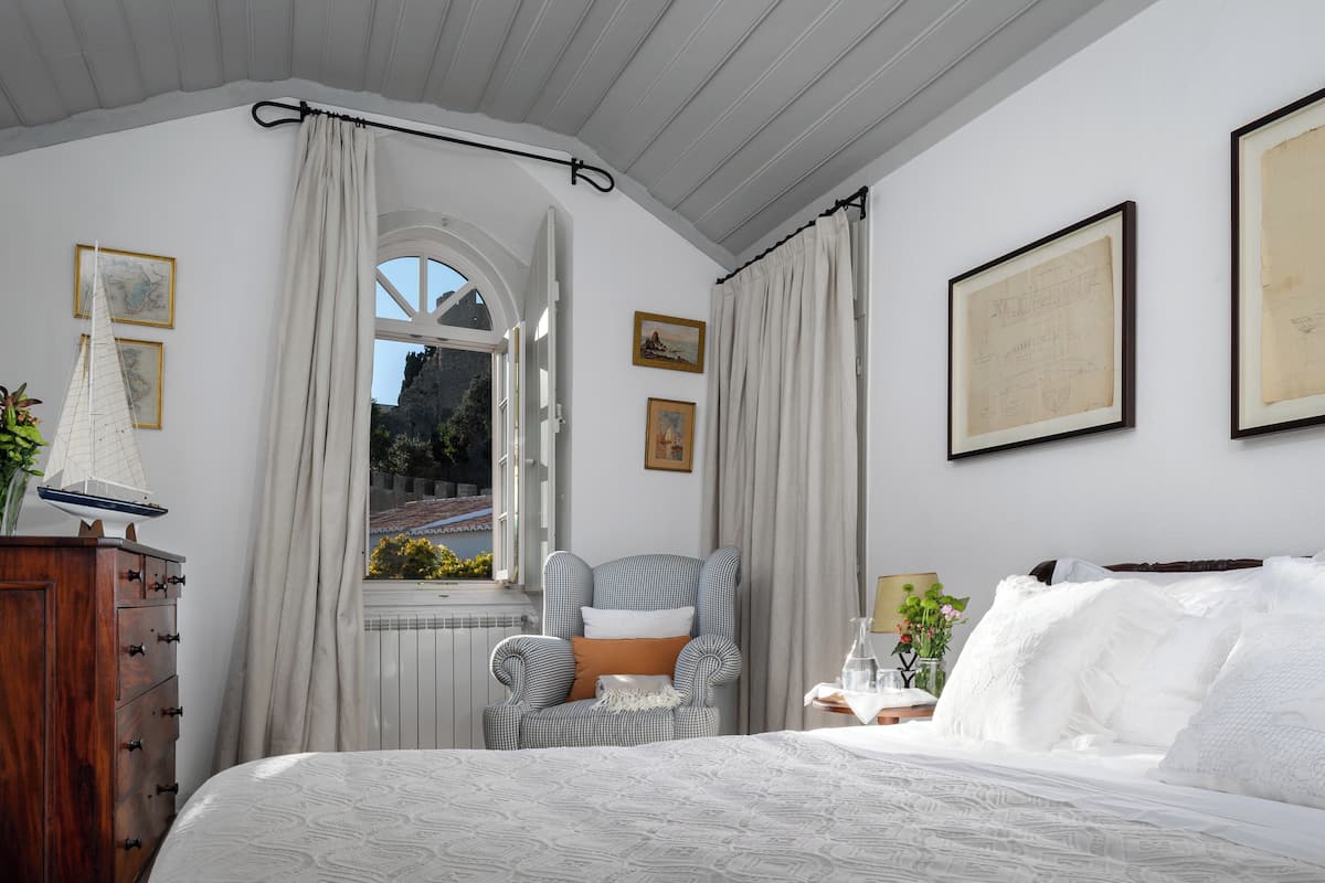 A bedroom with a large bed, a window with curtains, a chair, and artwork on the walls.
