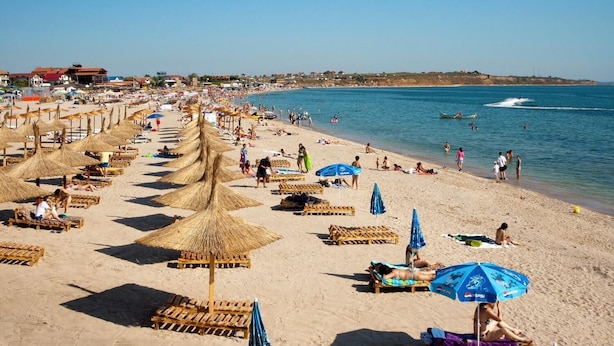Uma praia com fileiras de espreguiçadeiras e guarda-sóis, mar azul cristalino e edifícios ao fundo.