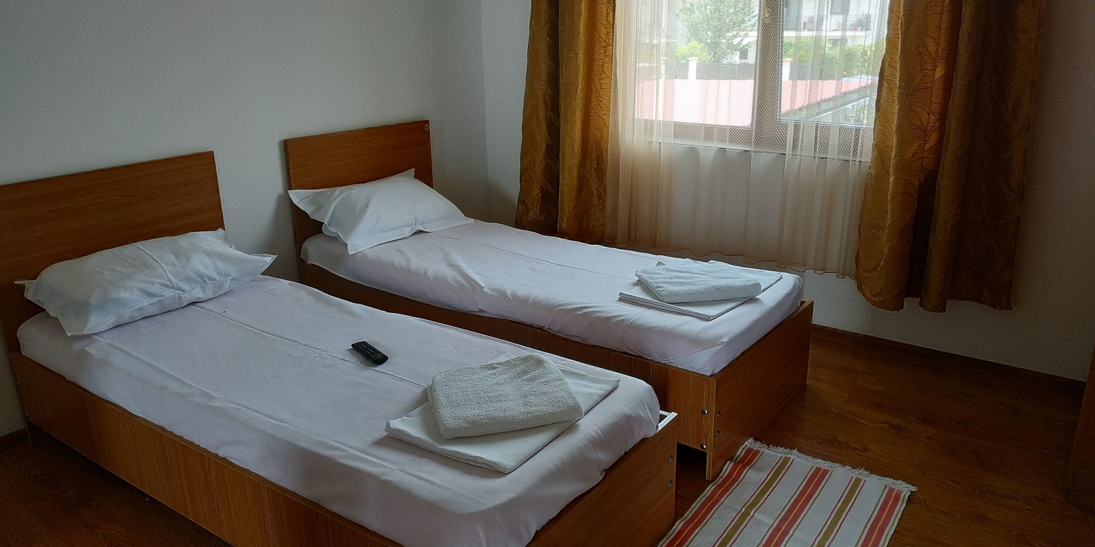 Deux lits simples avec des cadres en bois et du linge de lit blanc, une télécommande sur le lit de gauche et un tapis à rayures sur le sol.