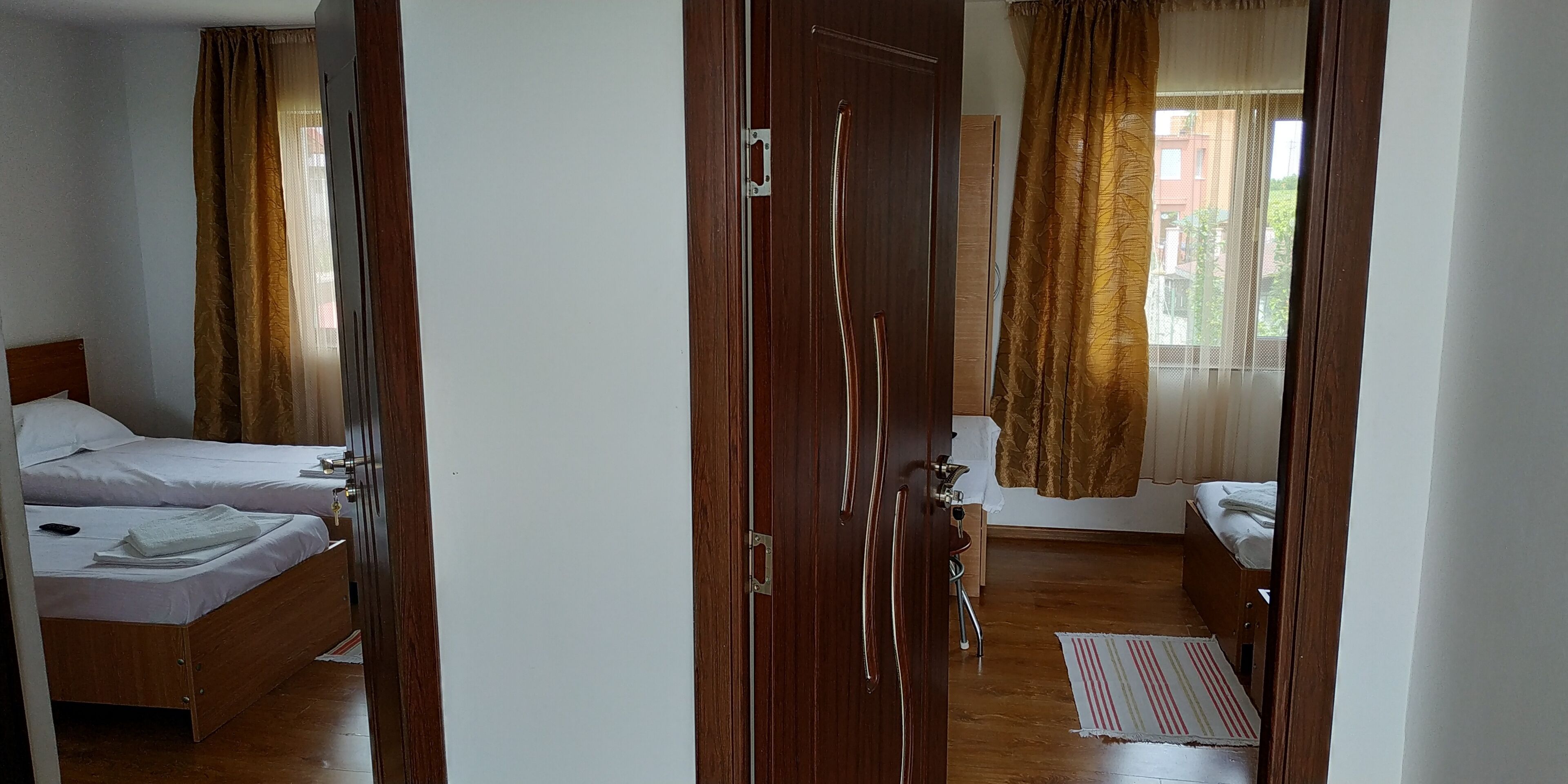A hotel room with two beds, wooden flooring, and a view of the outside through a window with curtains.
