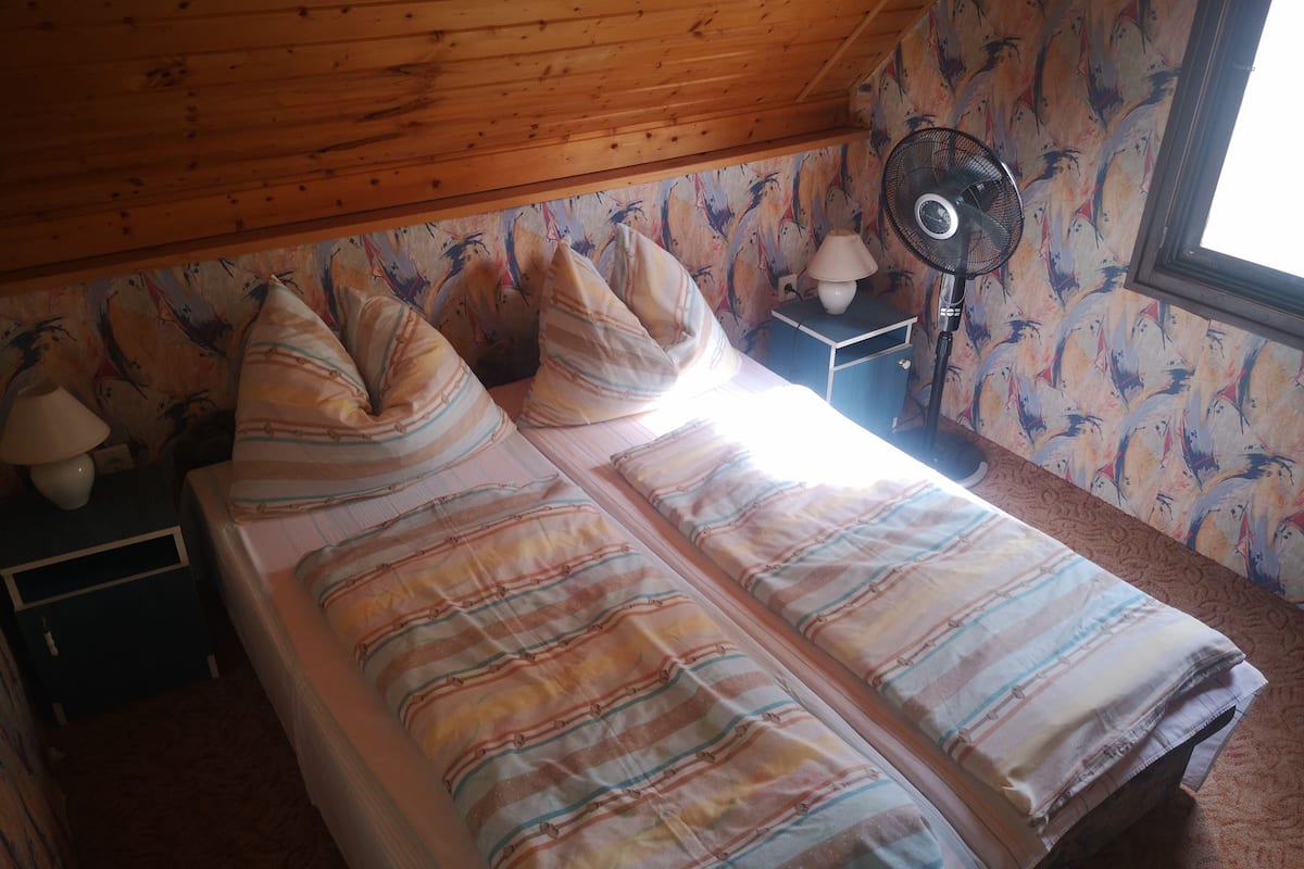 Un dormitorio con techo de madera, cama con ropa de cama a rayas, mesita de noche con lámpara y un ventilador de pared.