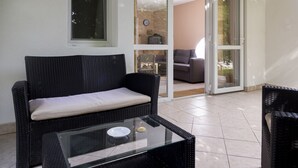 Un sofá de mimbre con un cojín, una mesa con superficie de vidrio que sostiene un plato y una cuchara, y un ventanal corredero que da a un espacio exterior con una silla y una mesa.