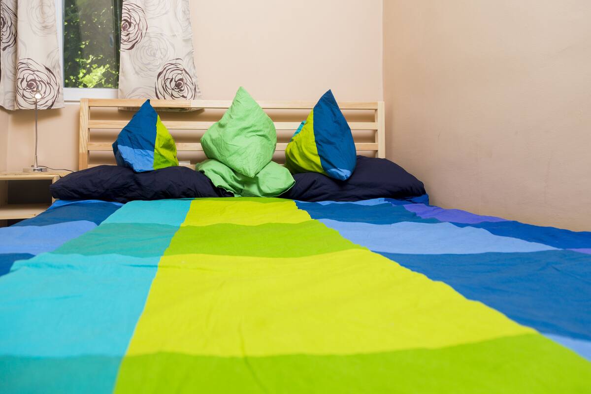 Una cama con colcha y almohadas a rayas coloridas, cabecera de madera y ventana con cortinas.