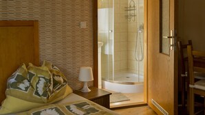Un dormitorio con techo de madera, una cama con ropa de cama floral, una mesita de noche con lámpara, y una puerta de madera que da a una zona de ducha.
