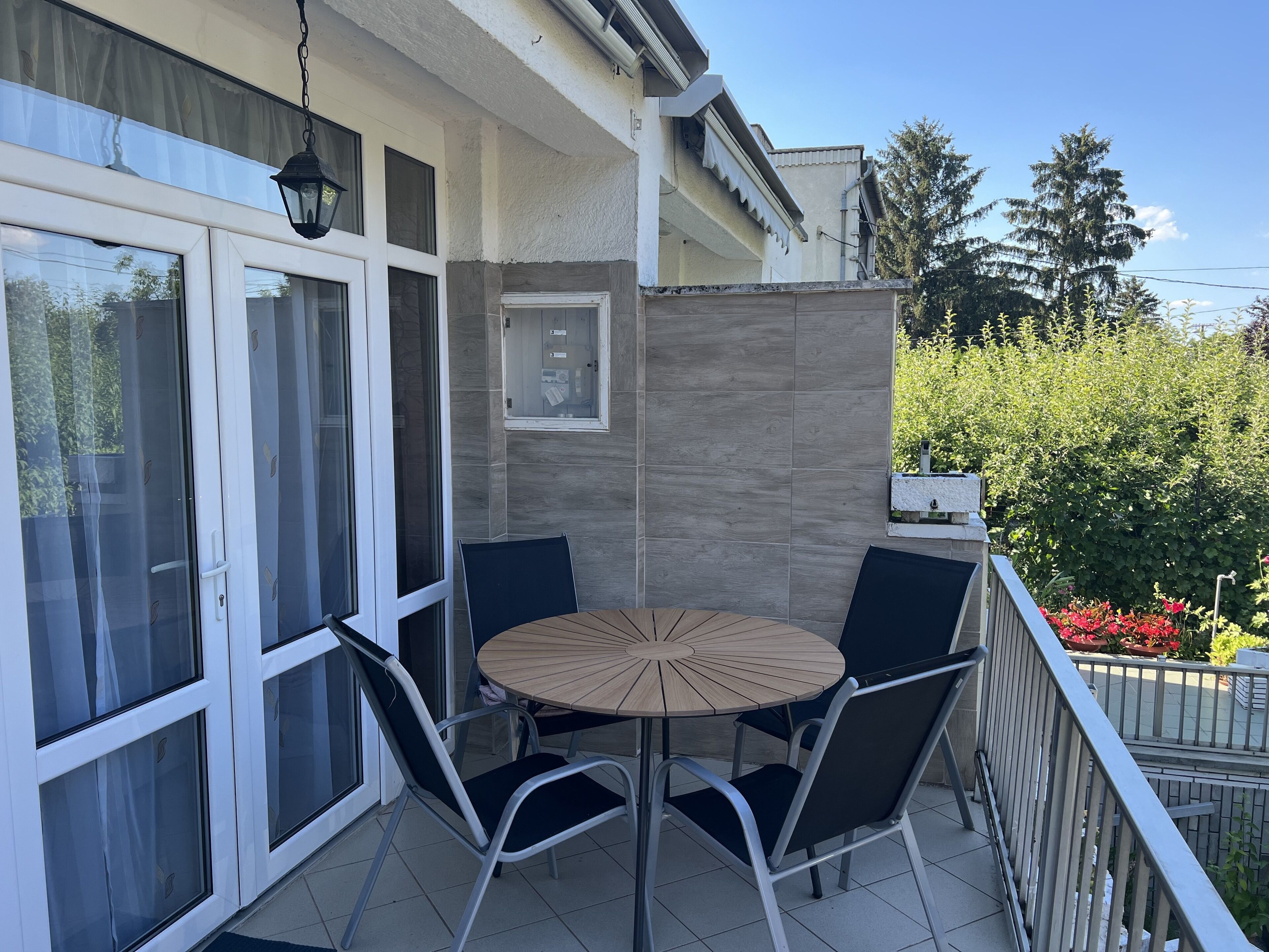 Ein Balkon mit rundem Tisch und vier Stühlen, eine Schiebeglastür und Blick auf Grünflächen sowie ein Gebäude gegenüber.