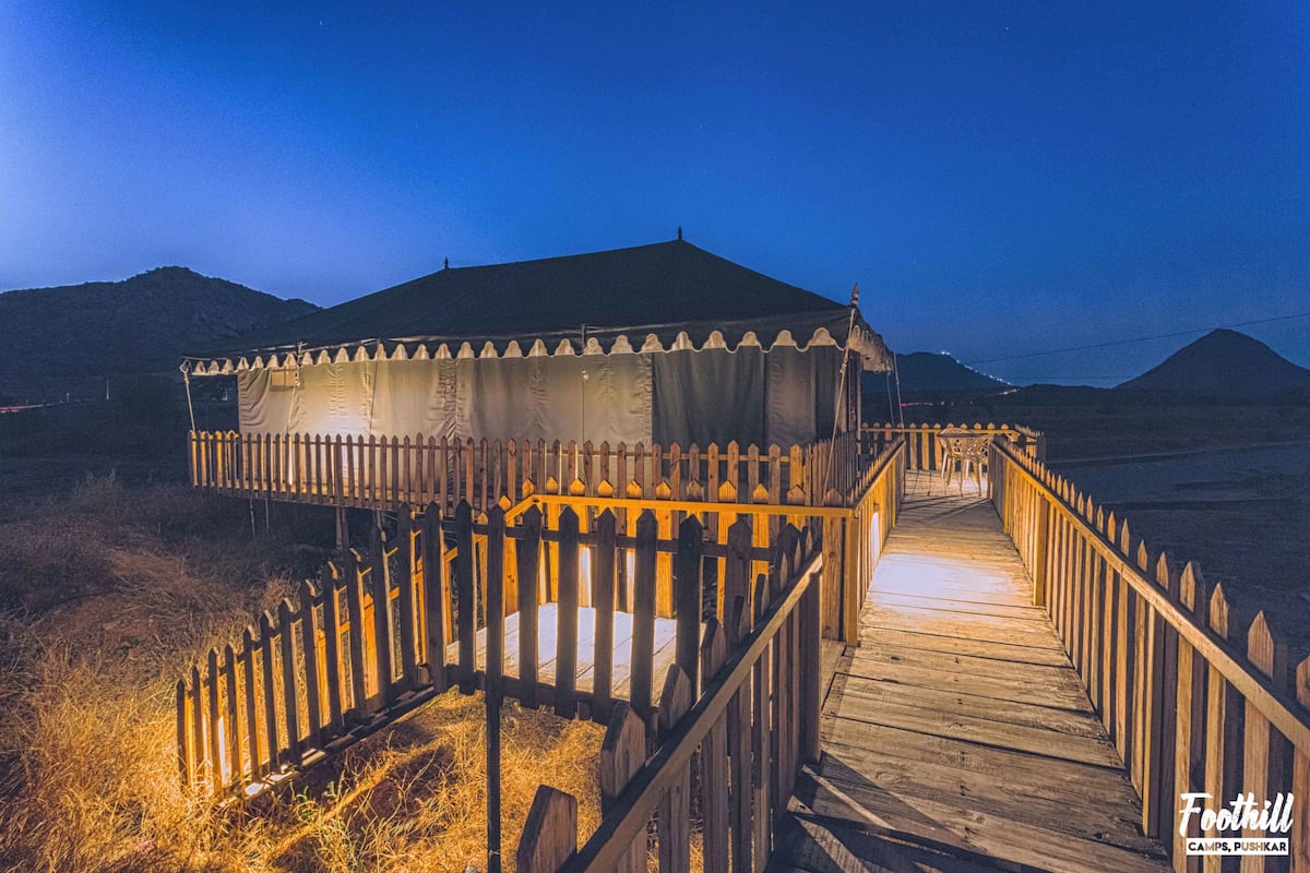 A wooden boardwalk leading to a tent-like structure with a fenced perimeter, set against a mountainous backdrop.