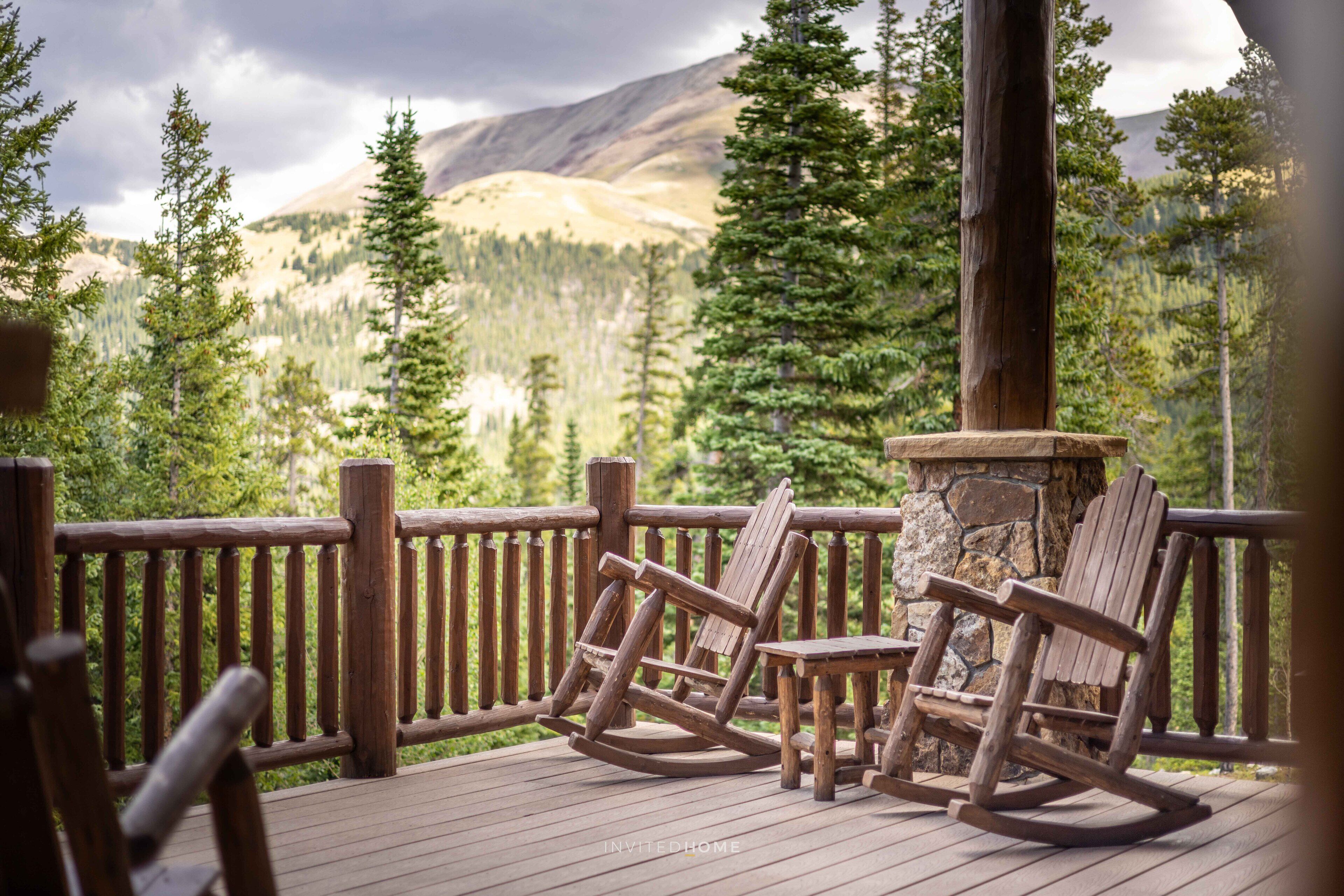 Une terrasse en bois avec deux fauteuils à bascule et une cheminée en pierre, offrant une vue sur une chaîne de montagnes boisée.