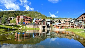 A lakeside resort with a central bridge, reflecting buildings, and a mountainous backdrop.