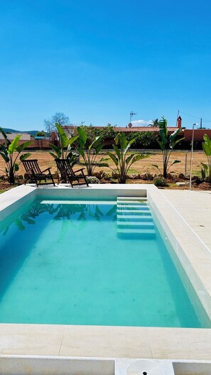 Ein rechteckiger Swimmingpool mit klarem, blauem Wasser, umgeben von einer gefliesten Terrasse. Zwei Liegestühle und mehrere Kübelpflanzen sind vorhanden. Im Hintergrund erstreckt sich ein klarer Himmel und im entfernten Bereich Gebäude.