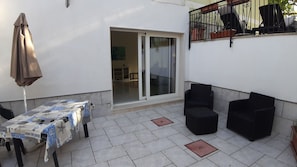 Een terras met een tafel, bedekt met een tafelkleed met een patroon, twee zwarte stoelen, een bruine parasol en een schuifdeur naar een binnenruimte.