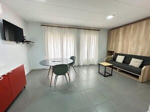 A modern living room with a round table, two chairs, a sofa, a TV mounted on the wall, and a window with curtains.