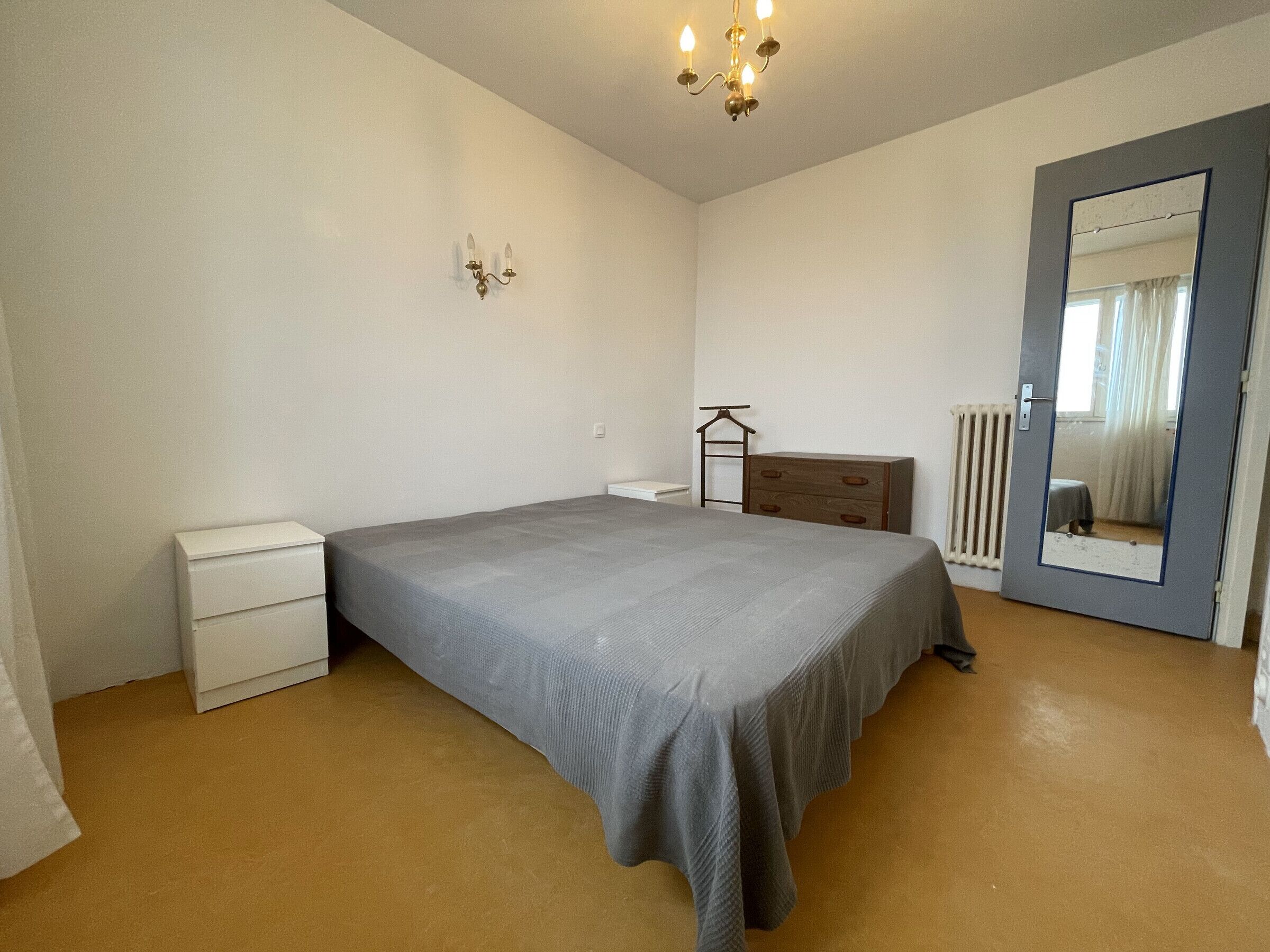 Une chambre à coucher comprenant un lit, une commode, une table de chevet, un miroir et un radiateur.