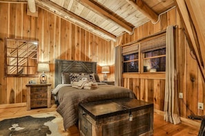 Ein Schlafzimmer einer Holzhütte mit einem Bett, Nachttischen, einer Kommode, einem Truhenkoffer und einem Fenster mit Vorhängen.