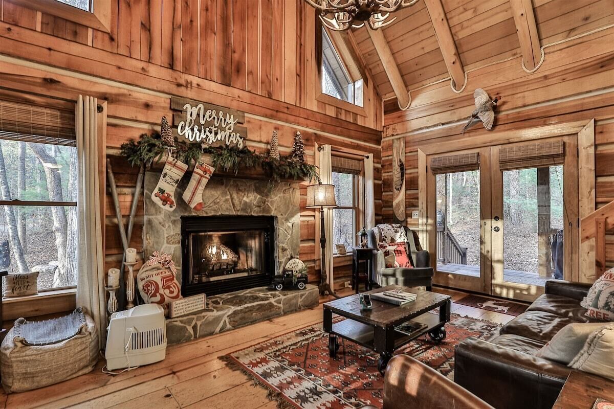Interior acogedor de una cabaña de troncos con chimenea, muebles de cuero y un letrero que dice 'Feliz Navidad'.