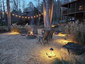 A rustic outdoor seating area with a fire pit, wooden chairs, and string lights.