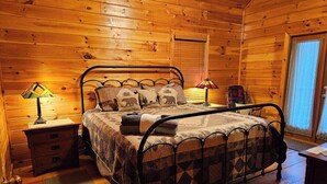A wooden cabin bedroom with a metal bed frame, two bedside tables with lamps, a window with curtains, and a door. - Casa Del Rio | Ellijay, GA (Ellijay)