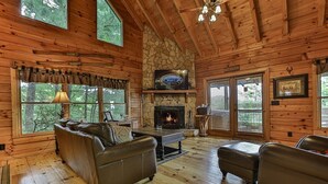 A log cabin interior with a stone fireplace, leather sofas, and large windows. - Moonlight Hideaway | Ellijay, GA (Ellijay)