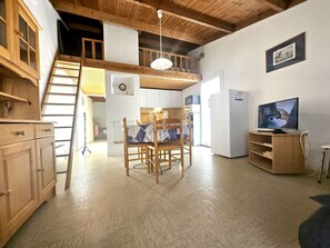Ein Loft-Zimmer mit Esstisch, Stühlen, Fernseher, Kühlschrank und einer Holztreppe.