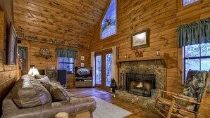 Ein Interieur einer Blockhütte mit Kamin aus Stein, Ledersofa, Schaukelstuhl aus Holz und einem Fernseher im Wohnzimmer.