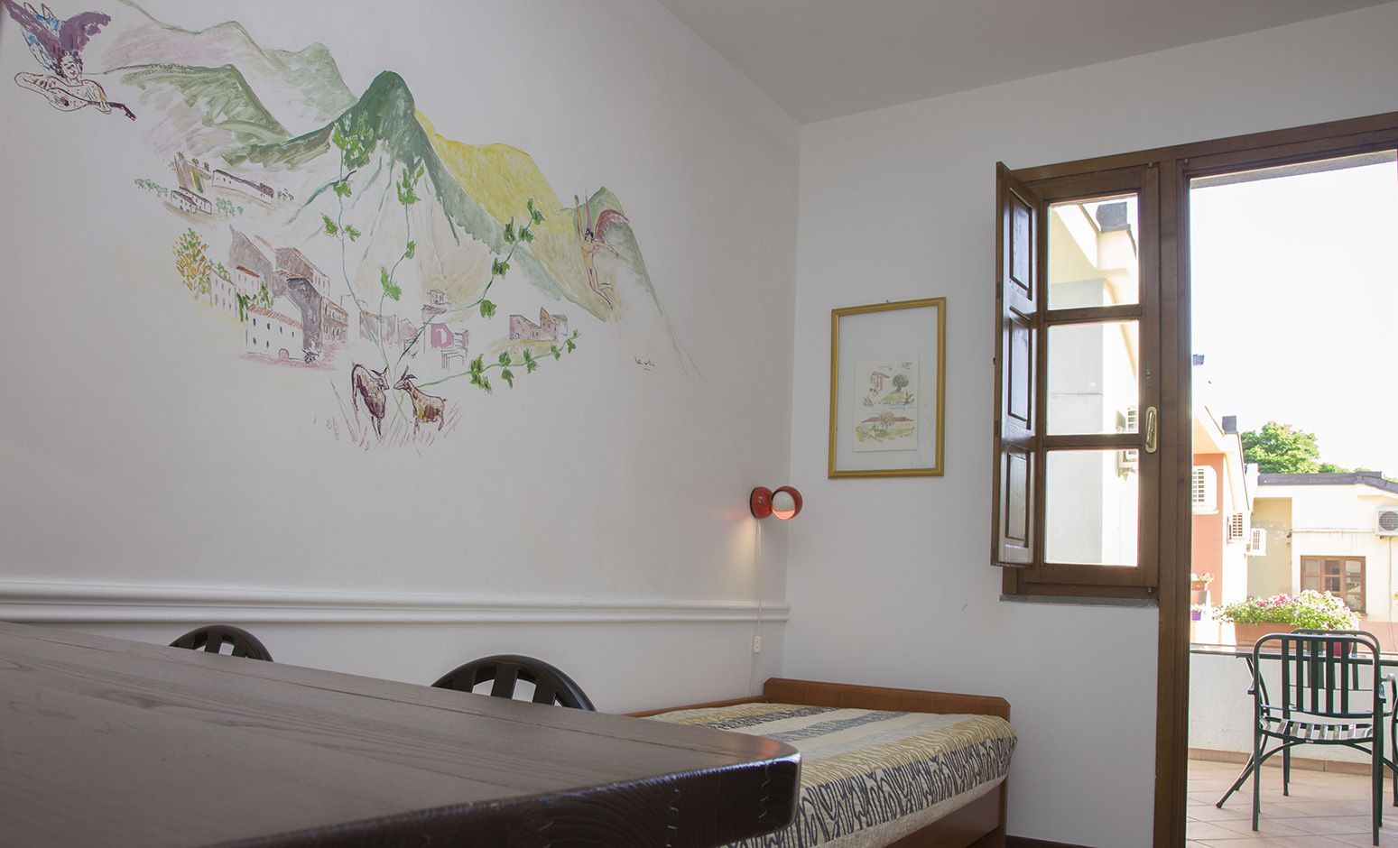 Una habitación con una mesa de madera, una cama con una colcha estampada, un mural en la pared, un cuadro enmarcado y una ventana con vistas a edificios.