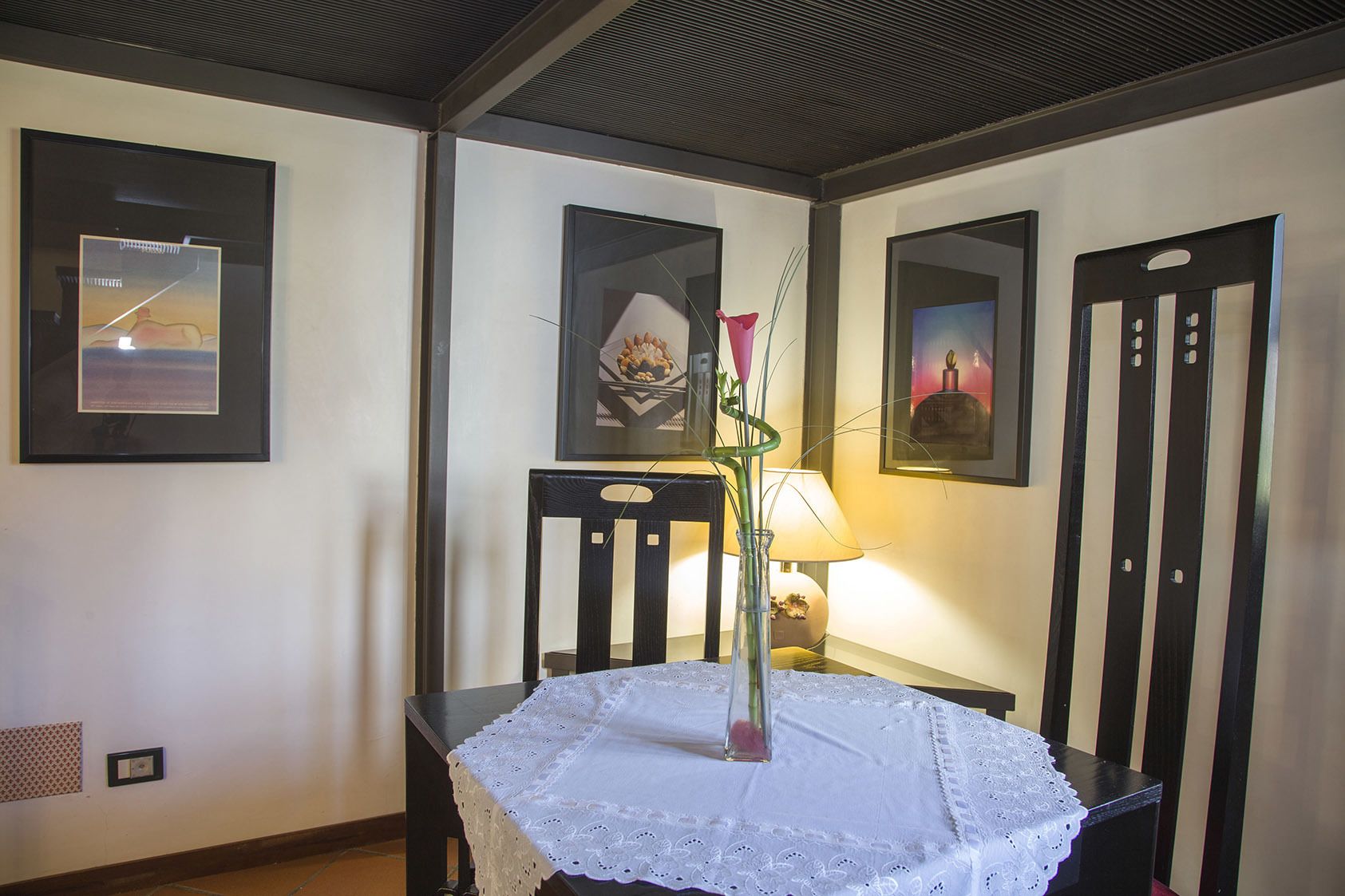 A dining area with a table covered by a lace tablecloth, framed artwork on the walls, and a vase with a pink flower.