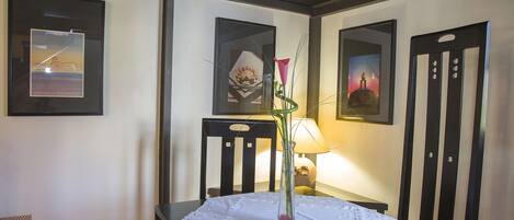 A dining area with a table covered by a lace tablecloth, framed artwork on the walls, and a vase with a pink flower.