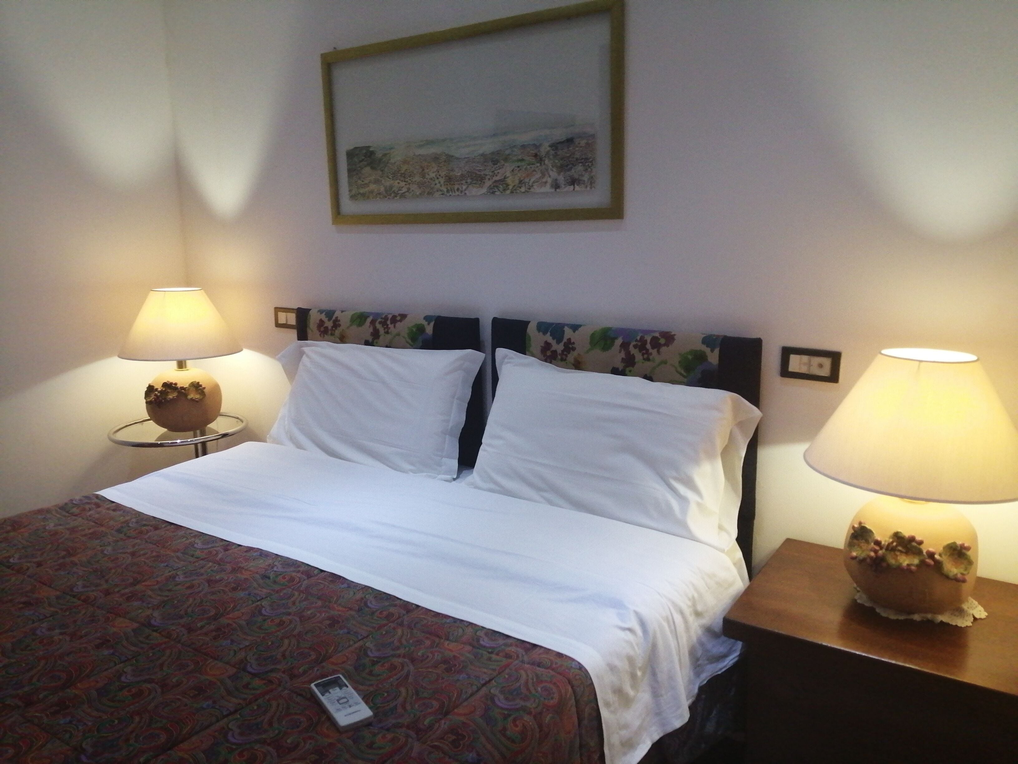 A neatly made bed with white linens, two bedside lamps, a framed picture on the wall, and a patterned carpet.