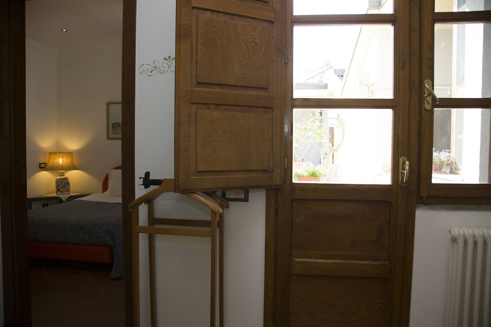 A bedroom with a bed, a nightstand with a lamp, a wooden wardrobe, and a window with a view of the outside.
