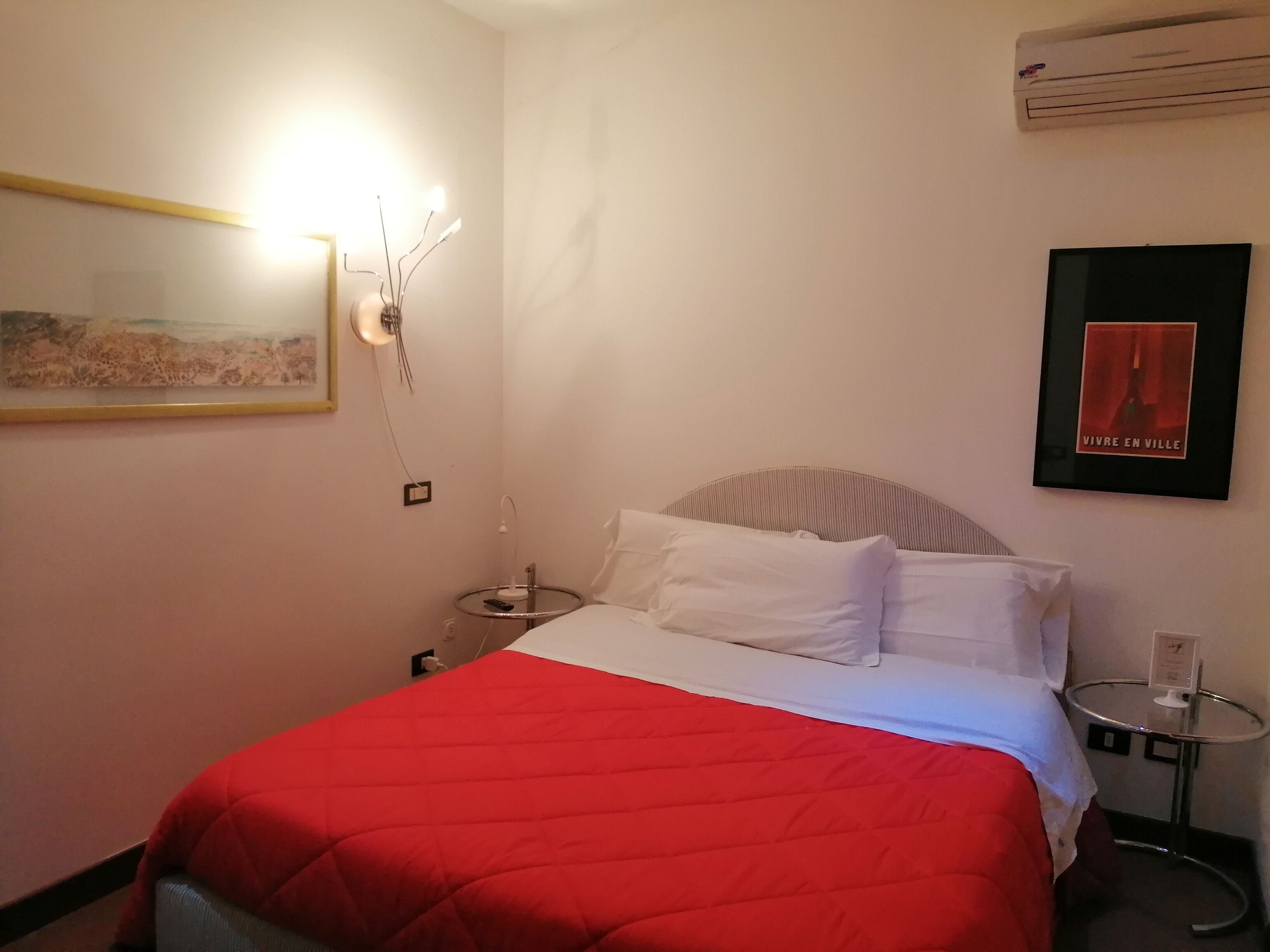 A hotel room with a bed, a bedside table, a wall-mounted air conditioner, a framed picture, and a wall-mounted light fixture.