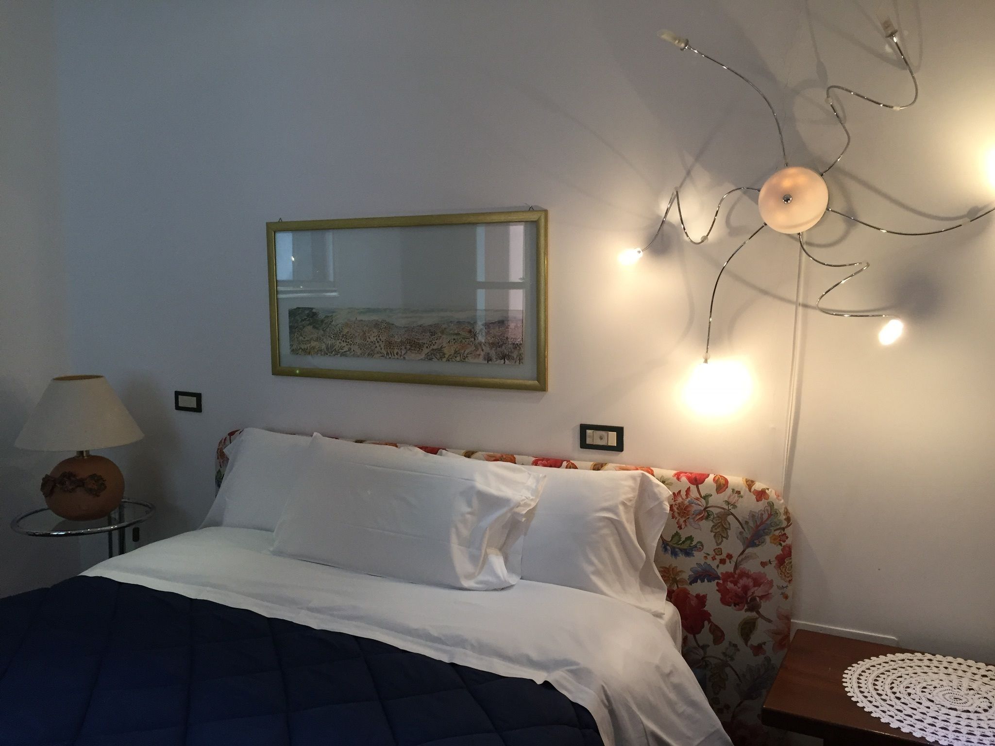 A bedroom with a bed, a nightstand, a lamp, a framed picture on the wall, and a ceiling light fixture.