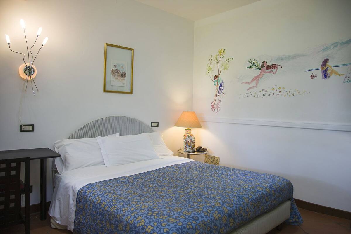 Un dormitorio con cama, mesita de noche, lámpara y una pared con mural.