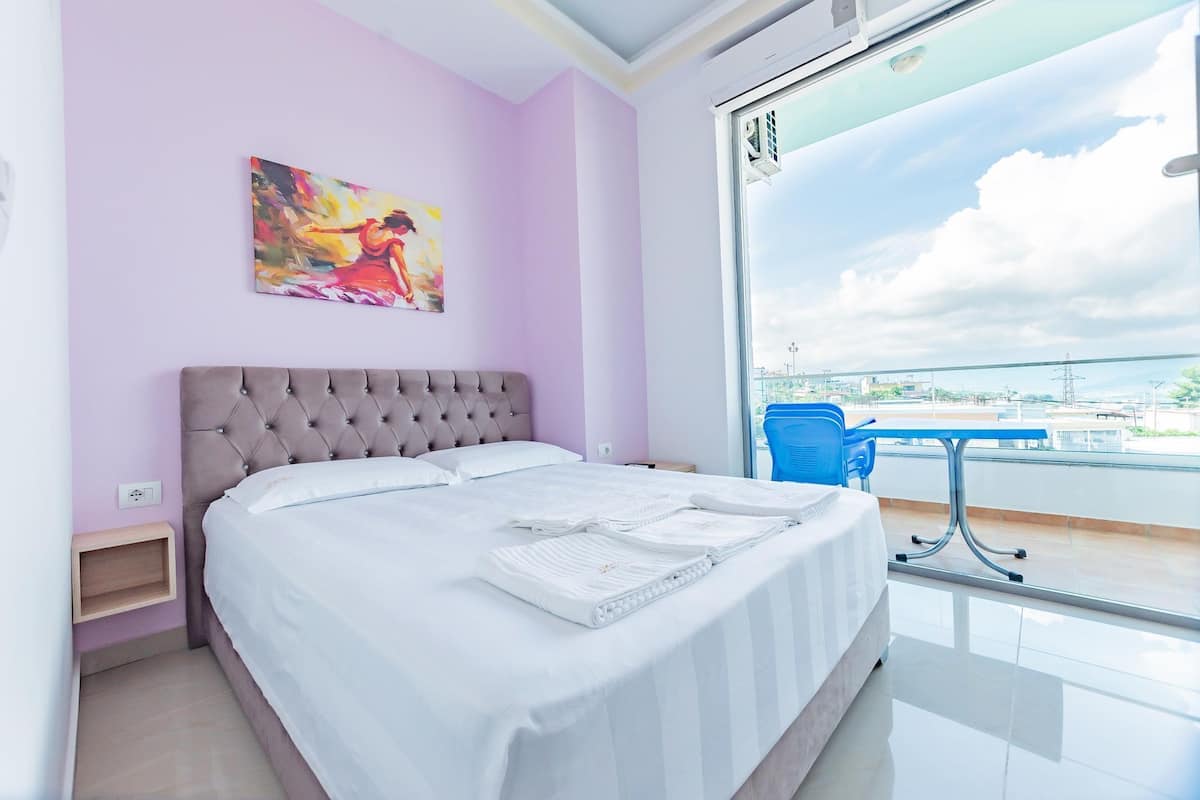 A modern hotel room with a large bed, a view of the city, and a balcony with a table and chairs.