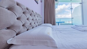 Et hotelværelse med en buet sengegavl, en stor seng med hvide sengetøj, udsigt over byen gennem en glasdør og et blåt bord ved vinduet.