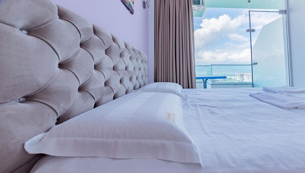 A hotel room with a tufted headboard, a large bed with white linens, a view of the city through a glass door, and a blue table by the window. - The City Gate Hotel (Sarandë)