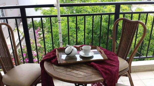 A balcony with wicker chairs, a small round table with a tray holding a teapot and cup, and a view of lush greenery. - Marysplacestalis (Hersonissos)