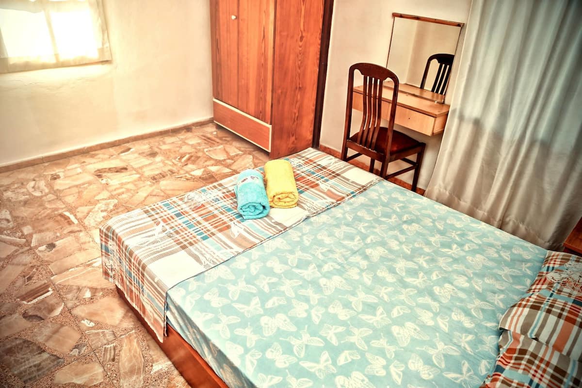 A bedroom with a bed, a wooden wardrobe, a desk with a chair, and a window with curtains.