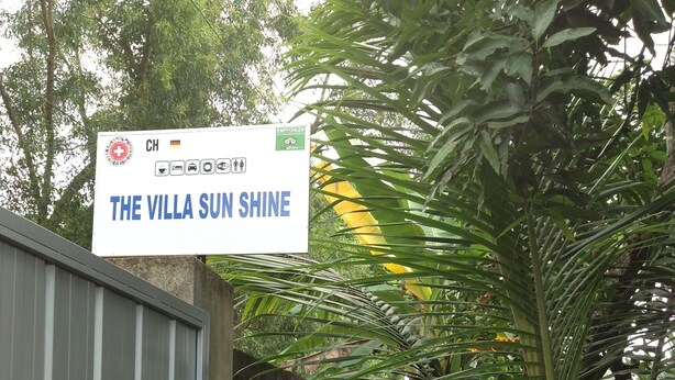 A sign for "The Villa Sun Shine" with a German flag, a medical cross, and an environmental symbol. - The villa sunshine (Divulapitiya)