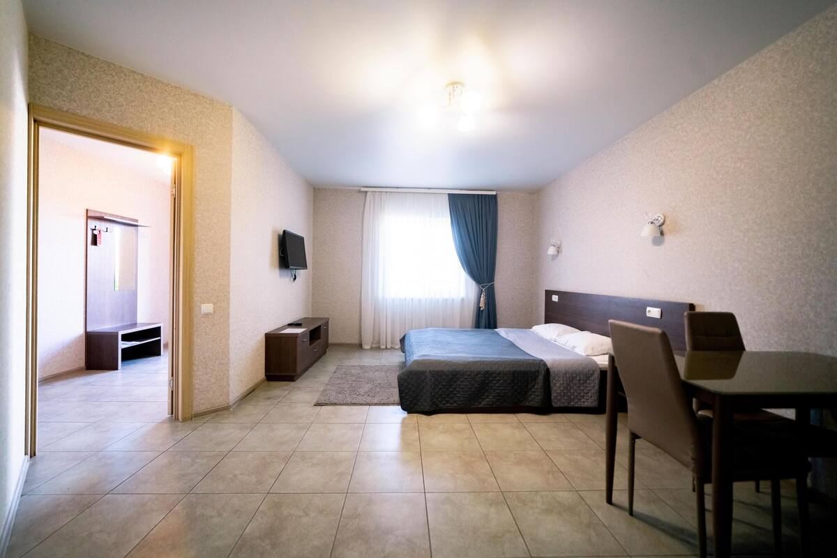 Quarto de hotel com cama, escrivaninha, cadeira, televisão e janela com cortinas.