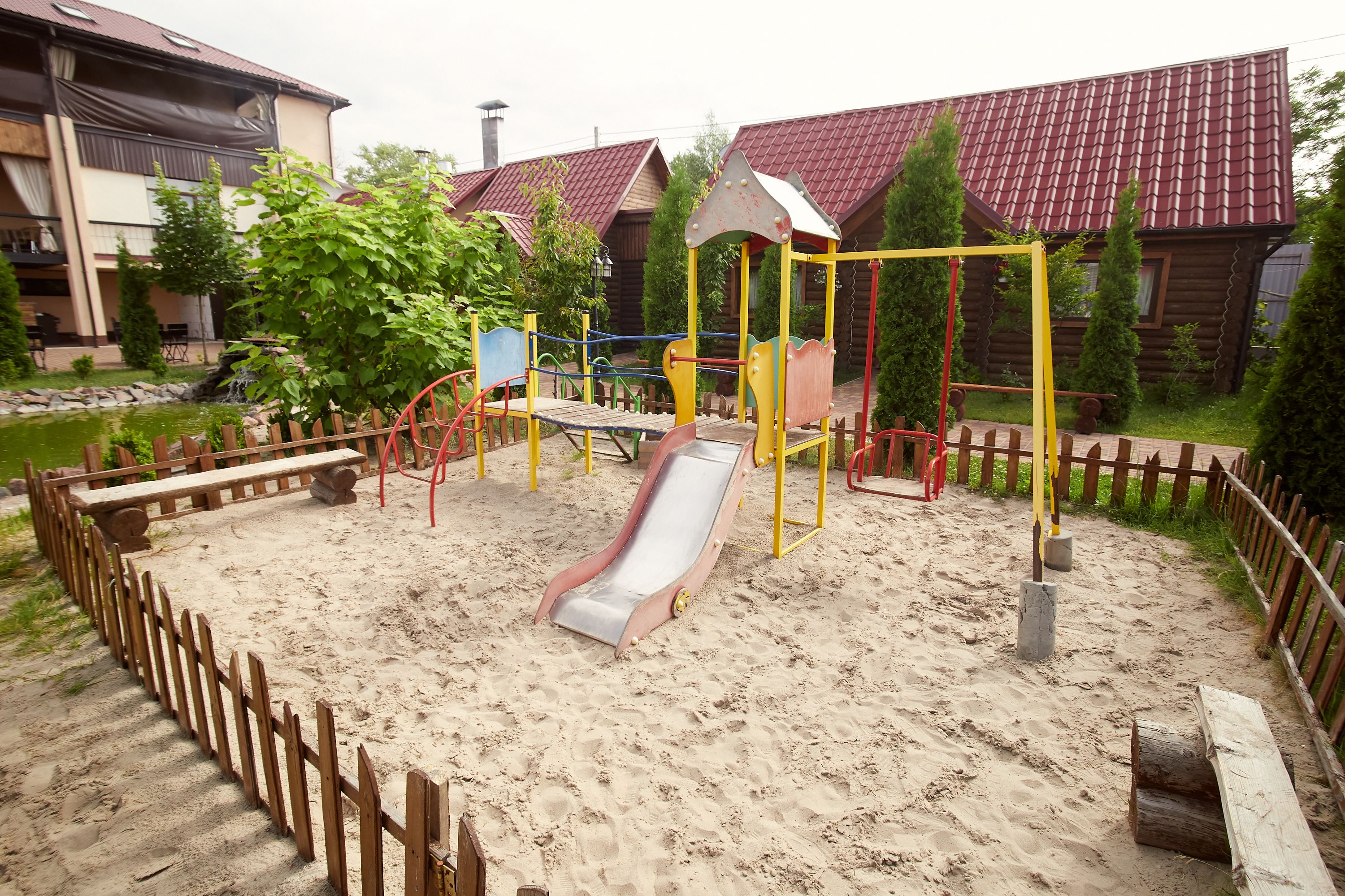 A playground with a slide and swings, surrounded by a wooden fence and sand.