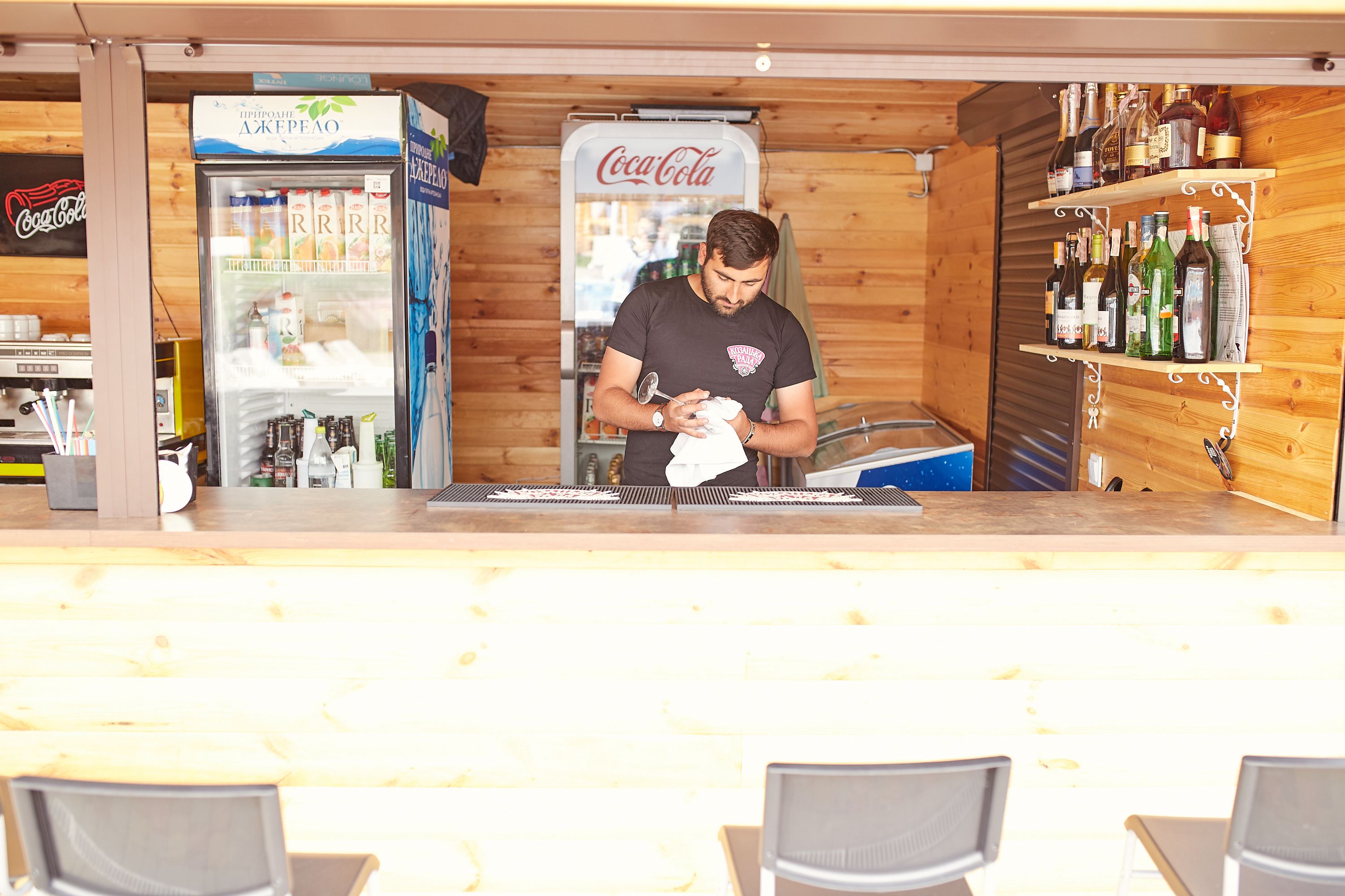 Un espace bar avec un réfrigérateur Coca-Cola, des étagères remplies de bouteilles et une personne qui prépare des boissons.