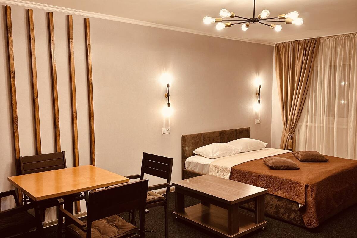Quarto de hotel com uma mesa de jantar de madeira, duas cadeiras, uma cama com cabeceira marrom, uma pequena mesa de cabeceira e um lustre no teto.