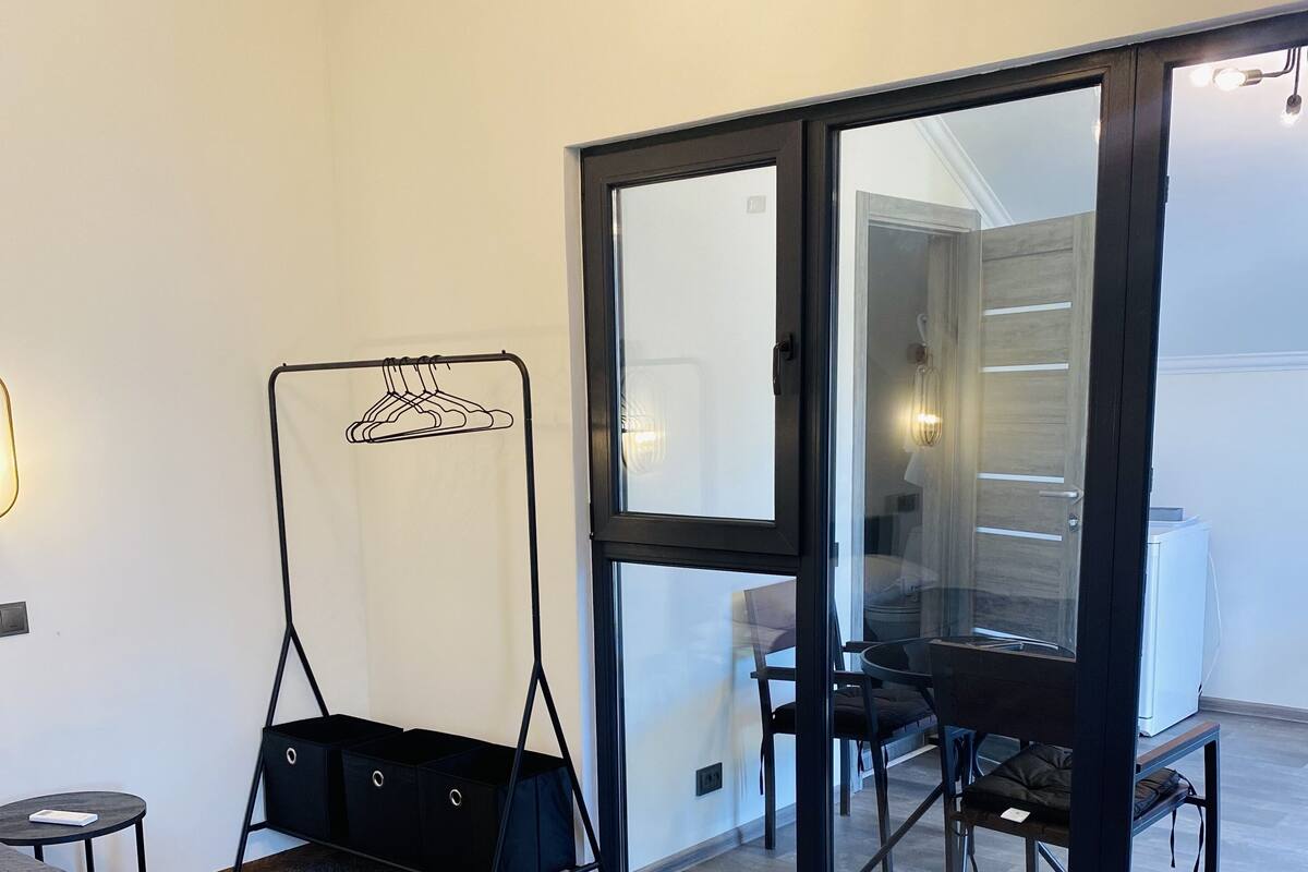 Quarto de hotel com cama, uma pequena mesa, uma cadeira, cabideiro e porta de vidro deslizante que dá para uma varanda com cadeiras.