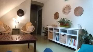 A cozy living room with a wooden table, two wicker chairs, a white bookshelf, and decorative wall art.