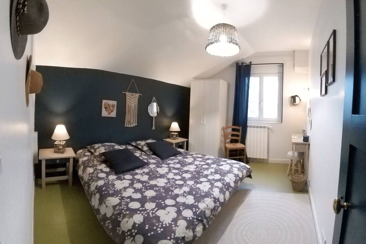 A bedroom with a bed, two bedside tables, a hanging light fixture, a chair, a wardrobe, and a window with curtains.