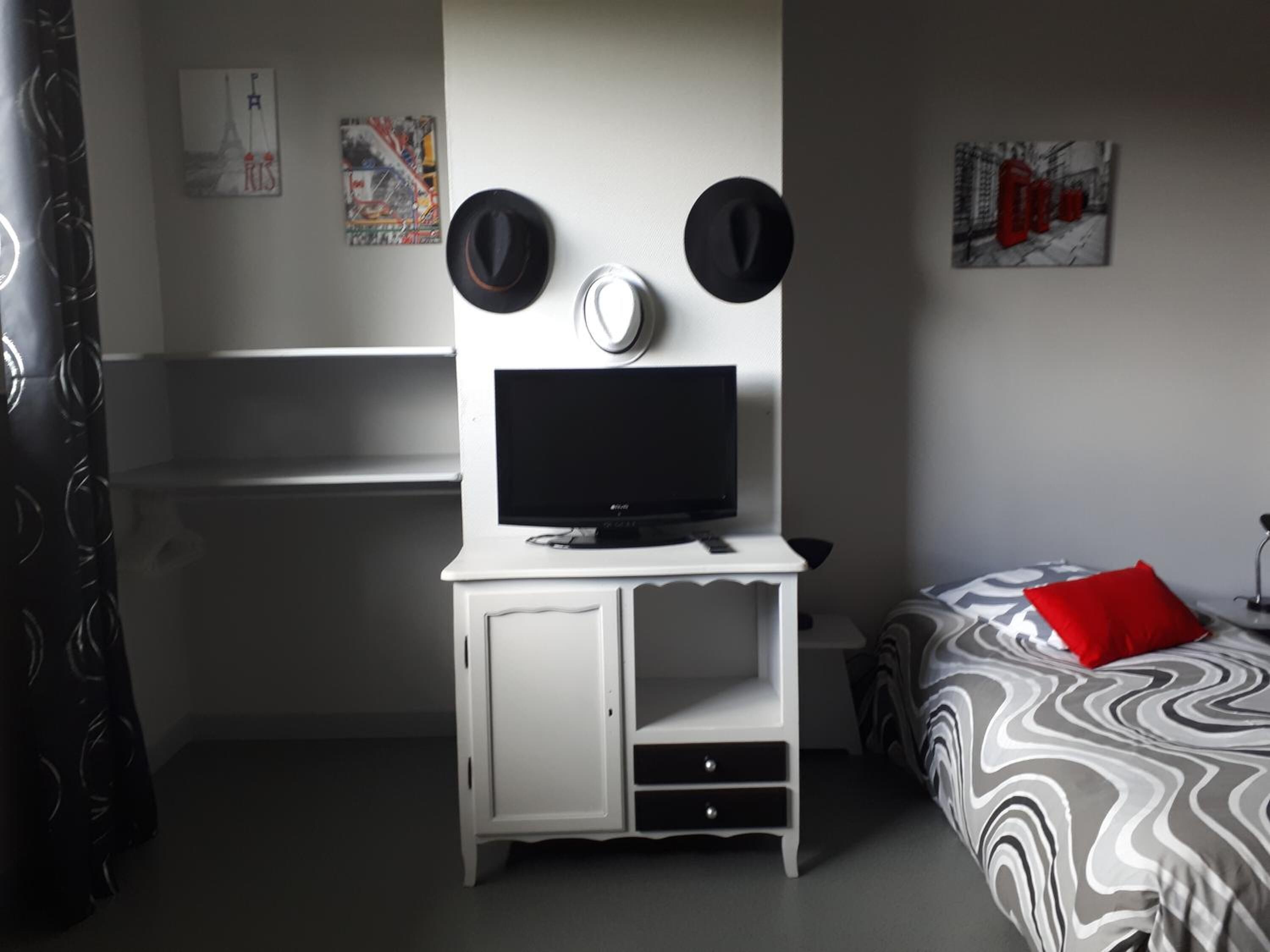 A bedroom with a TV on a white cabinet, a bed with a patterned blanket, and two hats on the wall.