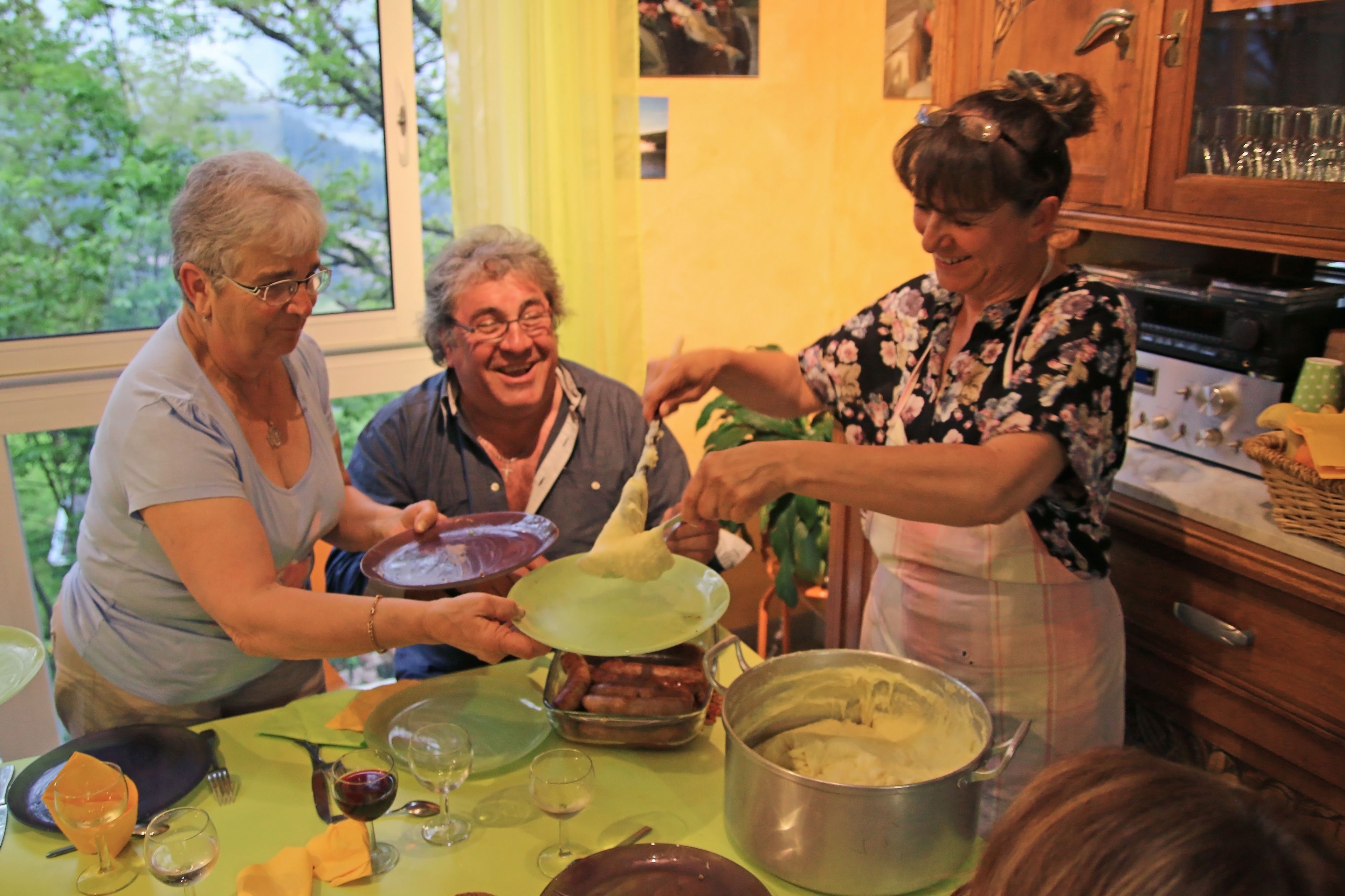 Un groupe de personnes se trouve dans une cuisine, en train de préparer des aliments. Une grande fenêtre est visible à l’arrière-plan.
