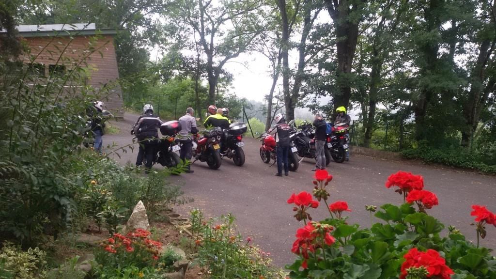 A group of motorcyclists gathered around motorcycles in a wooded area with a building in the background.